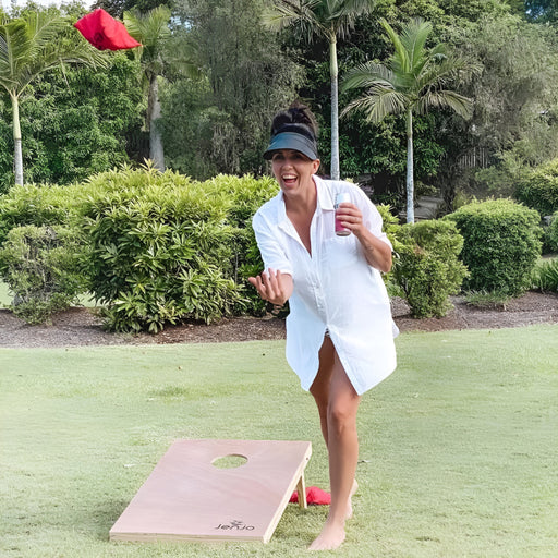 image of a lady wearing a hat holding a drink throwing a red bean bag with a cornhole board in the background with trees in the back and she is playing on grass