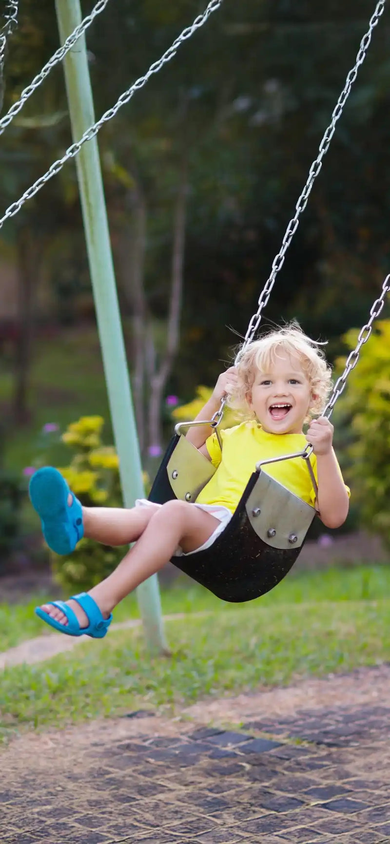 A joyful toddler with blonde hair wearing a yellow shirt and blue sandals, swinging on a black and grey swing.