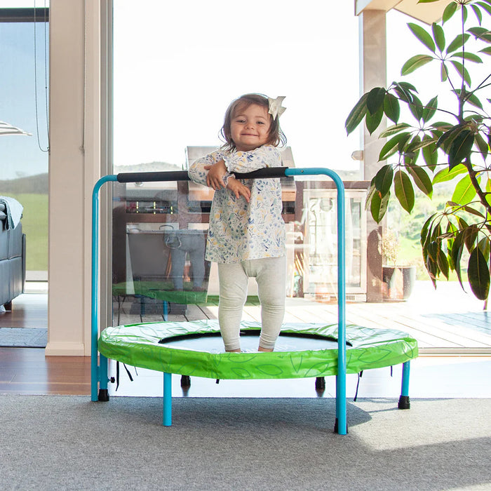 Child standing on a green and blue trampoline indoors with a plant and furniture in the background