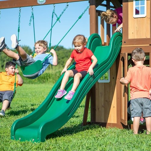 Close up image of 2 little boys swinging on the swings of Lakewood Swing And Play Set, a little girl sliding on the green slide, a little girl preparing to slide and a little boy entering the snack bar door
