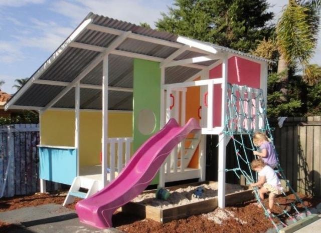 Mega Triplex Cubby House - painted in white;, yellow, pink, green and blue with little kids climbing on a climbing net and sandpit below