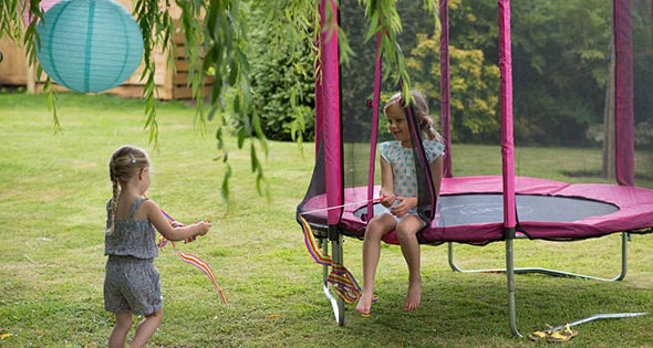 Half image of 4.5ft Junior Kids Trampoline Pink toddler trampoline on an outdoor background with 2 little girls, one sitting at the entrance of the trampoline and one standing outside the trampoline